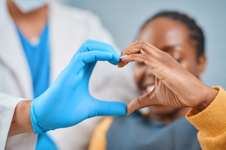 A healthcare professional in blue gloves forms a heart shape with their hands beside a smiling young Black patient, symbolizing trust, care, and positive dental experiences.