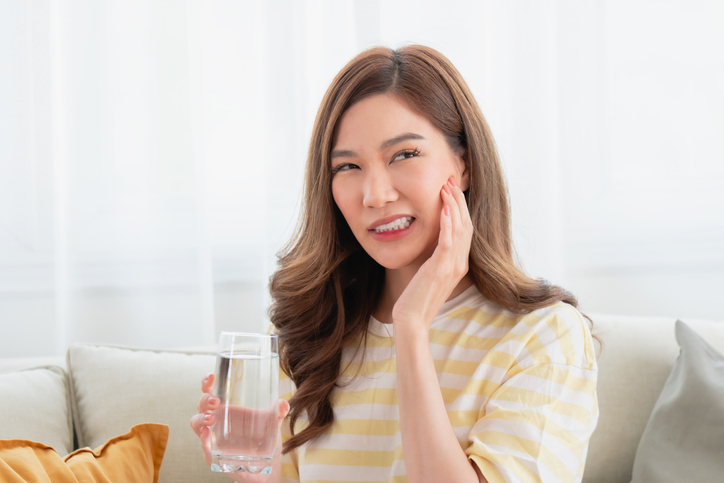 Young beautiful asian woman holding a glass of water and wincing in pain from tooth sensitivity or dental problems while sitting on a sofa at home.
